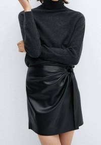 Jupe en cuir noir avec un design drapé et un détail noué à la taille, associée à un pull à col roulé gris foncé en maille douce.