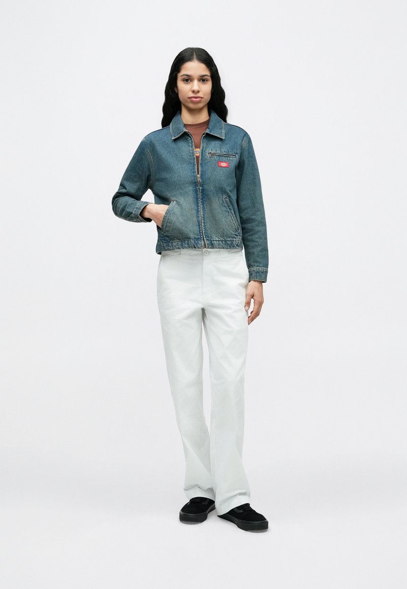 Jonge vrouw staand, gekleed in een blauwe spijkerjack, bruine top, witte broek en zwarte schoenen, tegen een effen witte achtergrond.