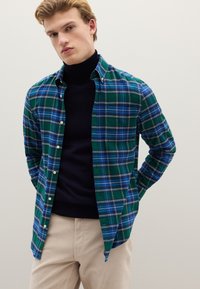 Camisa de cuadros verdes y azules con detalles en blanco, cuello abotonado, mangas largas y un ajuste relajado, combinada con un suéter de cuello alto negro debajo.