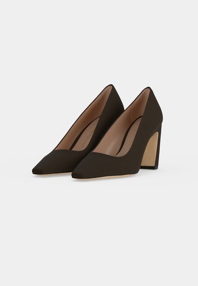 Une paire d’escarpins brun foncé à bout pointu et à hauts talons carrés, sur un fond clair.