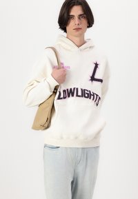 Felpa crema con ricami rosa e neri, con il testo "99 LYC YEARS" e "LOWLIGHTS". Indossata con jeans blu chiaro e una borsa a spalla color sabbia.