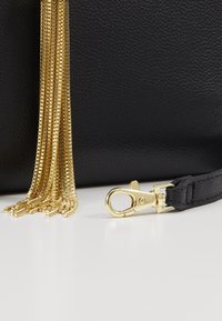 Musta nahkalaukku, jossa on teksturoitu pinta, kultaiset metallivetoketjut ja mustassa hihnassa oleva kiinnike.