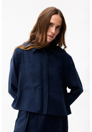 Femme aux longs cheveux bruns portant une chemise ample bleu foncé à boutons et un pantalon assorti, debout devant un fond blanc.