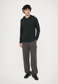 Capuz de malha em verde escuro com cordão, combinado com calças cargo cinzas de corte largo. O conjunto apresenta um design texturizado.