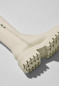 Bota impermeable de color crema con suela de goma texturizada, cremallera decorativa y un acabado liso. Cuenta con un diseño de suela gruesa para mayor tracción.