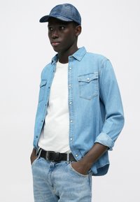 Gaiši zilas džinsa krekls ar pogu priekšpusi un divām krūšu kabatām, valkāts virs baltas T-kreklu, kopā ar gaiši zilām džinsu biksēm un rakstainu zilu cepuri.