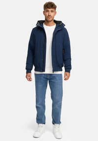 Chaqueta acolchada azul marino con capucha y cierre de cremallera, con forro negro y bolsillos laterales. Combinada con una camiseta blanca y jeans de mezclilla.