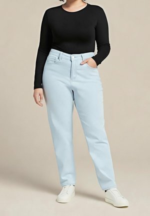 Mujer vestida con un top negro de manga larga, vaqueros de talle alto de color azul claro, zapatillas blancas, con una mano en el bolsillo, de pie sobre un fondo beige.