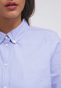 Chemise à boutons bleu clair avec une texture lisse, col pointu et boutons blancs le long du devant, affichant un design décontracté.
