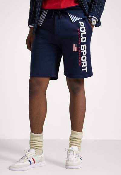 Marineblå treningsshorts med elastisk midje, utstyrt med "POLO SPORT" og et amerikansk flagg-design i rødt og hvitt. Kombinert med hvite sko.