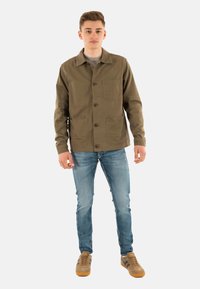 Veste en coton vert olive avec un col, quatre poches avant, fermeture à boutons, portée sur un t-shirt gris et un jean bleu clair.