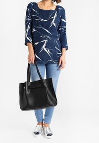 Borsa tote nera in pelle con forma strutturata, manici doppi, superficie liscia, posizionata accanto a una persona che indossa una maglietta stampata blu marina e jeans blu.