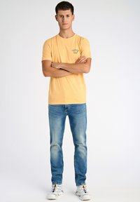 Camiseta de algodón de manga corta en un amarilllo claro con un pequeño gráfico en el pecho, combinada con jeans de mezclilla azules y zapatillas blancas.