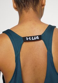 Teal urheilu-trikoo, jossa on urheilullinen takakappale. Varustettu mustalla hihnalla, jossa on "UA"-logo, sekä teksturoidulla kankaalla hengittävyyden takaamiseksi.