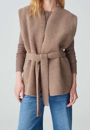 Femme portant une veste sans manches en polaire teddy marron clair ceinturée, par-dessus un haut taupe à manches longues et un jean bleu, debout, les bras détendus.