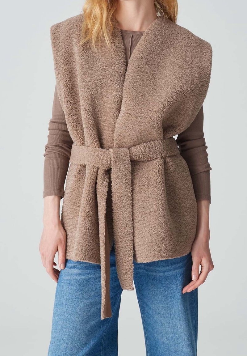Femme portant une veste sans manches en polaire teddy marron clair ceinturée, par-dessus un haut taupe à manches longues et un jean bleu, debout, les bras détendus.