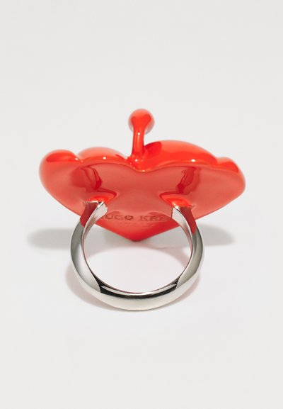 Red glossy heart-shaped accessory with a silver metallic ring. The heart features a protruding stem and embossed branding on the back.