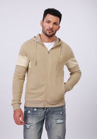Beige zip-up hoodie met crèmekleurige accenten op de mouwen, kangoeroezak, geribbelde manchetten en een gouden embleem op de borst. Draagt met versleten jeans.