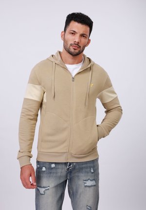 Beige zip-up hooded sweatshirt with cream accent sleeves, kangaroo pocket, ribbed cuffs, and gold emblem on the chest. Worn with distressed jeans.