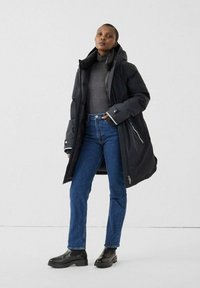 Vrouw staat in een zwart gewatteerd jack over een grijze coltrui, blauwe jeans en zwarte enkellaarsjes tegen een effen witte achtergrond.