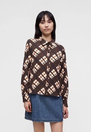 Femme portant une chemise à carreaux marron et beige à manches longues avec une jupe en jean bleue, debout devant un fond uni.