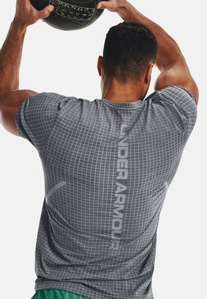 Mann in grauem Under Armour-Shirt hält einen Medizinball über dem Kopf und bereitet sich auf eine Übung vor, von hinten vor einfachem Hintergrund gesehen.