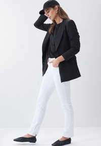 Veste noire à double boutonnage sur un top noir, associée à un jean blanc slim et des chaussures noires à enfiler. Casquette noire et accessoires minimalistes.