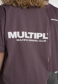Lilla bomulds-T-shirt med fed hvid tekst "MULTIPLY" og "MULTIPLY APPAREL CLUB" på ryggen. Korte ærmer og afslappet pasform.