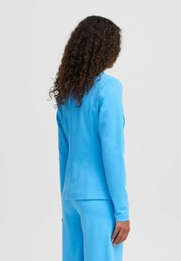 Blauwe, langwerpige jas met een getailleerd ontwerp. Glad materiaal, zichtbare naden en een hoge halslijn. Bijpassende blauwe broek is inbegrepen.