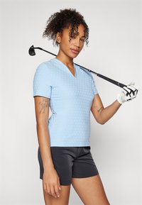 Polo bleu clair à manches courtes avec un col en V et une texture à pois, associé à un short noir. Un club de golf repose sur l'épaule.