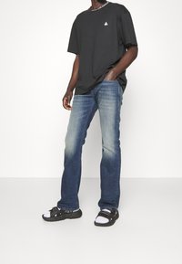 Musta puuvillainen t-paita, jossa on pyöreä kaula-aukko ja valkoinen logo, yhdistettynä sinisiin farkkuihin, joissa on haalistunut muotoilu. Mustat ja valkoiset urheil sandaalit.