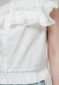 Weiße Baumwollbluse mit Rüschensleevs, Spitzenverzierung und Knopfverschluss. Sie weist ein strukturiertes Muster entlang der Brust und einen fransigen Saum auf.