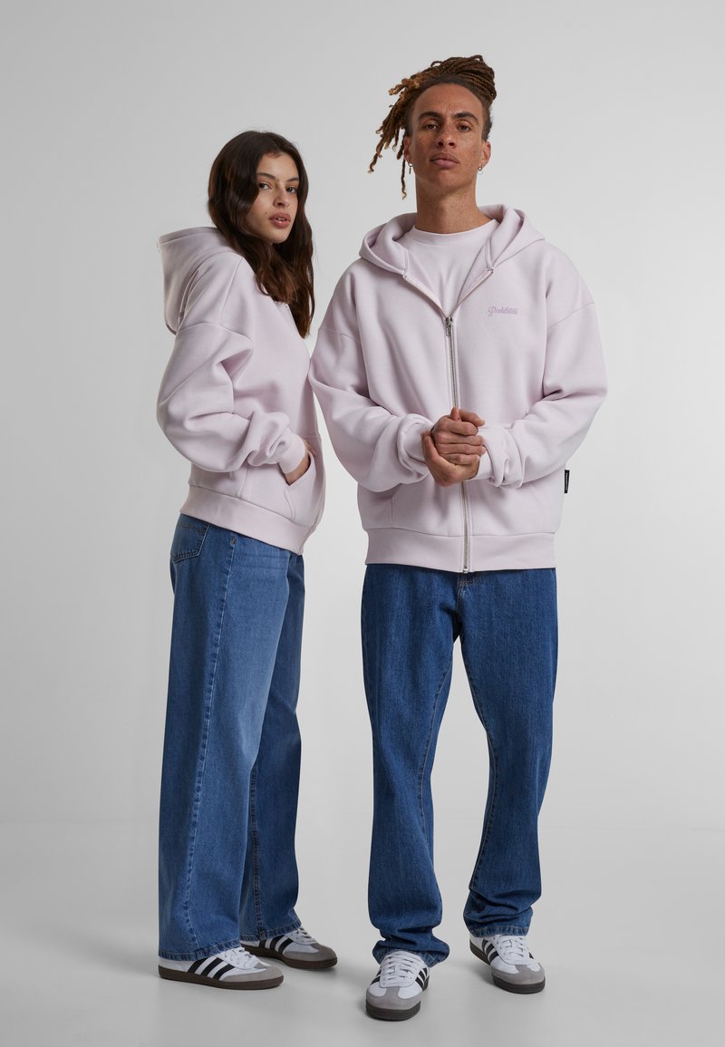Felpa zip-up rosa chiaro con un ampio cappuccio, polsini a coste e un piccolo logo. Abbinata a jeans in denim blu e sneakers casual.