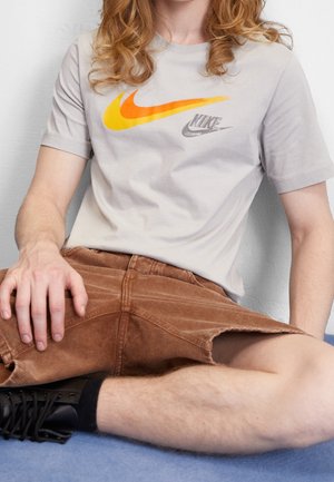 Jasnoszara bawełniana koszulka z pomarańczowo-żółtym logo Nike. W zestawie brązowe sztruksowe szorty o luźnym kroju z widocznymi szwami.