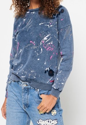 Personne portant un sweat-shirt bleu avec des motifs de tâches de peinture blanches, roses et noires et un jean bleu clair avec des patchs et de la broderie.