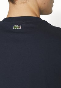 Man wearing navy blue shirt with small green crocodile logo on upper back, facing away against white background.