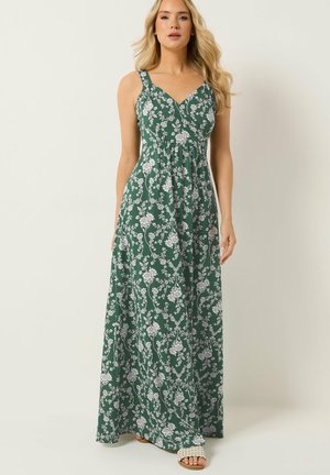 Vrouw draagt een groene mouwloze maxi-jurk met witte bloemen en beige open teen sandalen, staand tegen een effen achtergrond.