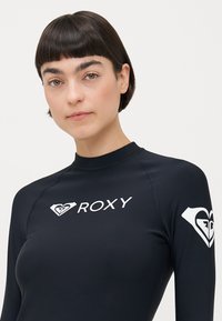 Crni dugorukavi rash guard s bijelim Roxy logotipom na prsima i lijevom rukavu. Značajke uključuju visoki ovratnik i mekan, glatki materijal.