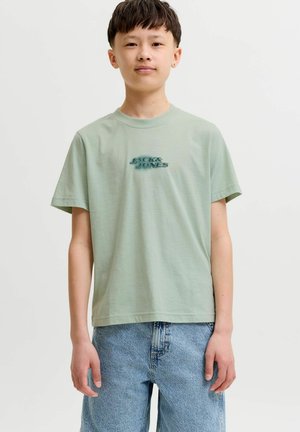 Jong persoon draagt een lichtgroen "Jack & Jones" t-shirt en blauwe spijkerbroek, staand tegen een effen witte achtergrond.