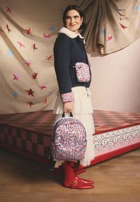 Femeie în jachetă bleumarin cu mansete și guler cu imprimeu floral, fustă crem în straturi, ținând un rucsac floral și purtând o borsetă asortată și pantofi roșii.