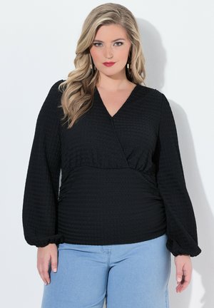 Vrouw met lang blond haar die een zwarte, getextureerde V-halsblouse met lange ballonmouwen en lichtblauwe spijkerbroek draagt, tegen een witte achtergrond.