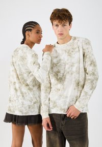 Koszula z długim rękawem w tie-dye w kolorze białym i oliwkowym; cechuje się teksturowanym materiałem, luźnym krojem oraz okrągłym dekoltem.