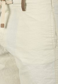 Pantalones cortos de lino en beige claro, con una textura suave, cintura ajustable con cordón y acento de cuero, y bolsillos laterales, diseñados para la comodidad.
