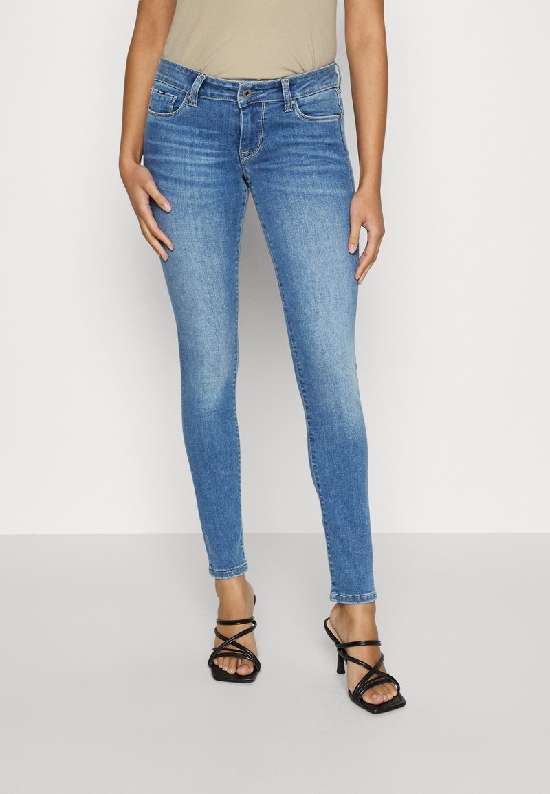 Jeans ajustados de mezclilla azul con cintura media, bolsillos frontales y textura suave, combinados con sandalias negras de tacón alto con tiras.