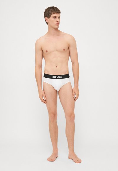 Biancheria intima bianca con una fascia nera che presenta il logo "VERSACE". L'elastico in vita è elastico. Il modello è in piedi a piedi nudi, mostrando un fisico snello.