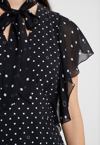 Černé šaty s bílými puntíky, které mají průhledné, lehké krátké rukávy a zavázaný detail na krku. Hladká textilie, elegantní design.