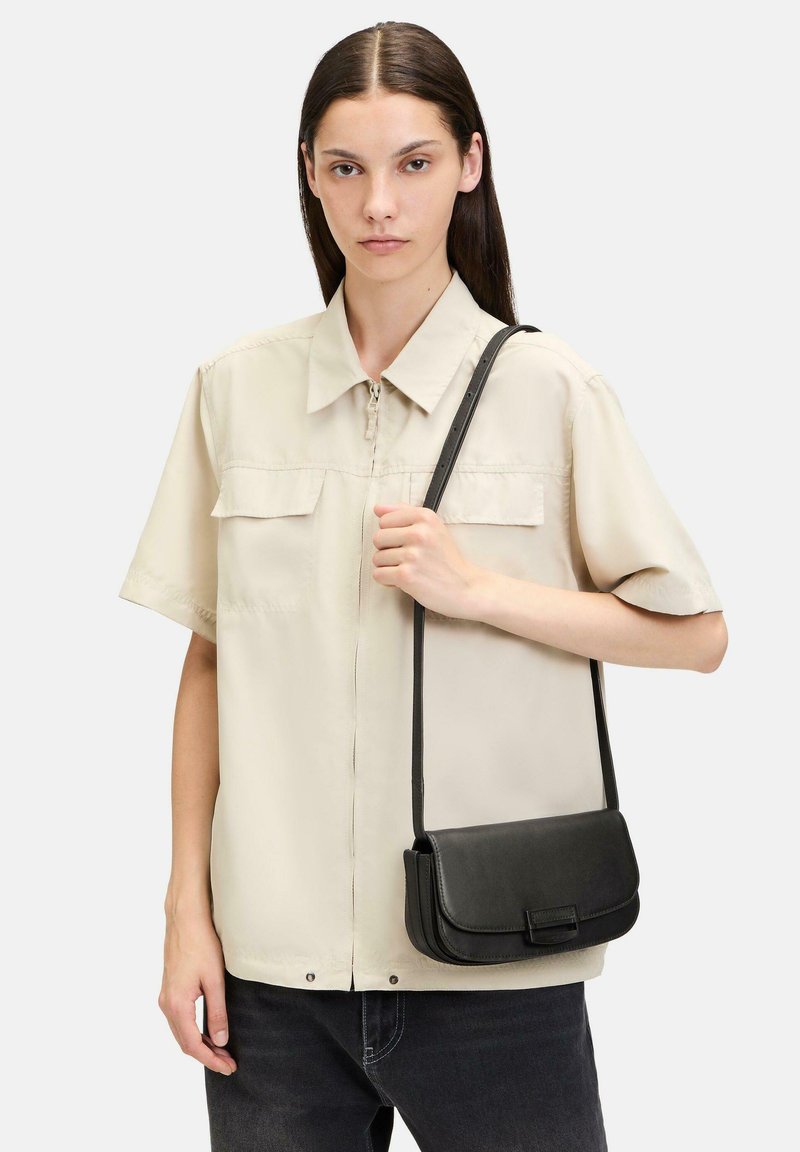 Camicia beige a maniche corte con chiusura a zip e due tasche frontali, abbinata a una piccola borsa a tracolla nera, indossata sopra jeans scuri.