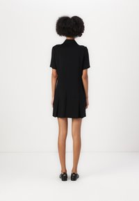 Abito nero a maniche corte con colletto e dettagli plissettati sull'orlo, realizzato in tessuto liscio. Indossato con scarpe nere.