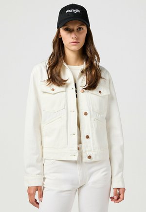 Weiße Jeansjacke mit Brusttaschen und Kupferknöpfen, kombiniert mit einem schlichten weißen Unterhemd und weißen Hosen, ergänzt durch eine schwarze Mütze.