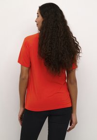 Raudona trumpa rankovė t-shirt, lygus paviršius, laisva vejimė, apvalus kraštas, dėvima su juodomis kelnėmis. Matomi ilgi, garbanoti plaukai.